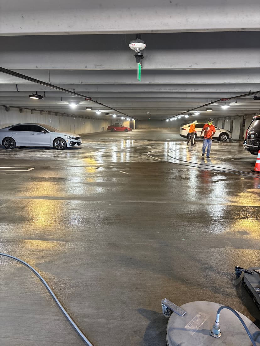 Cleaned parking garage with workers, cars, and reflective wet floor lights. Safety cones visible.